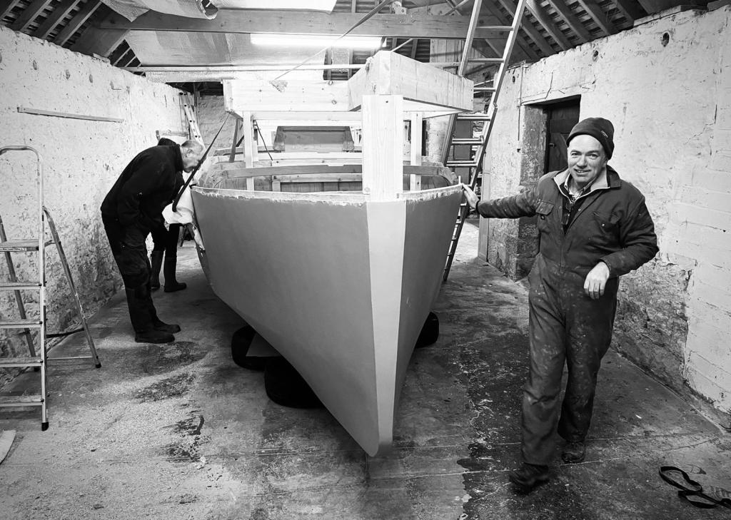 black and white photo of man smiling wearing working overalls and hat as he rests his hand on half a boat being built