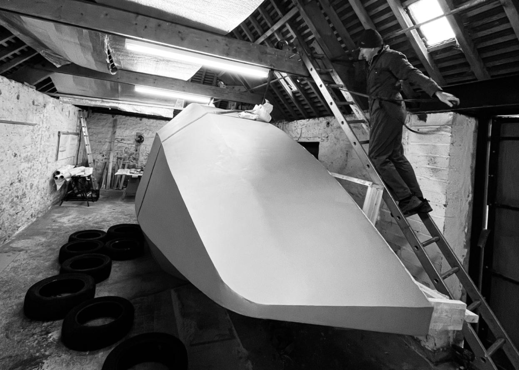 black and white photo of a man in overalls and hat standing on a ladder looking over a boat turned on it's side